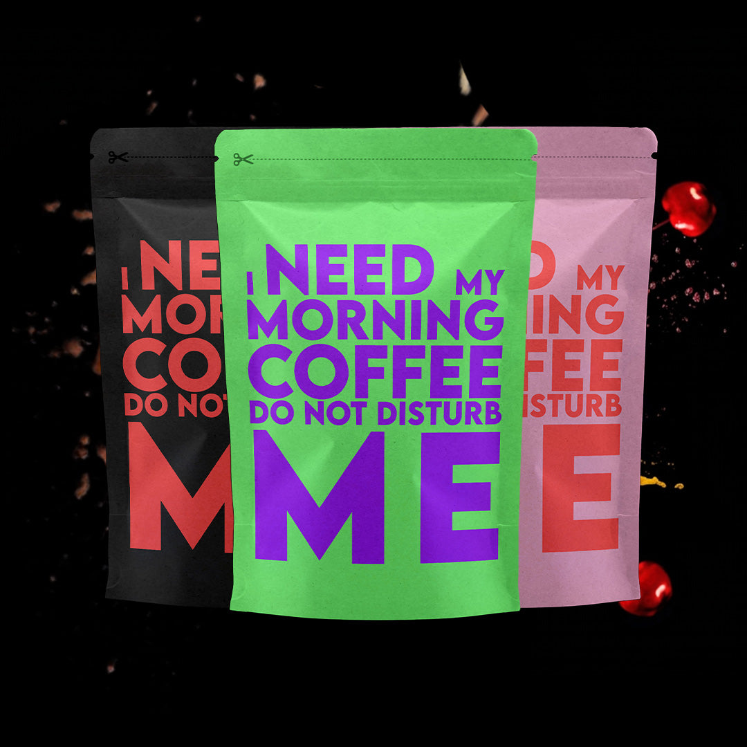 Drie koffiezakken met de tekst "I need my morning coffee. Do not disturb me", in verschillende kleuren: zwart met rode tekst, groen met paarse tekst en roze met rode tekst, tegen een donkere achtergrond met zwevende koffie-elementen en spetters.