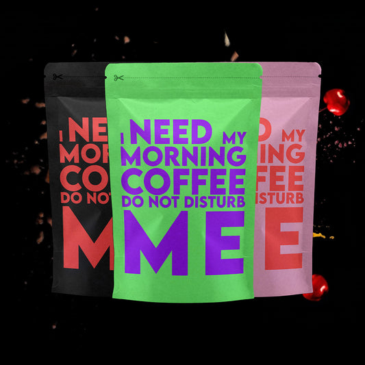 Drie koffiezakken met de tekst "I need my morning coffee. Do not disturb me", in verschillende kleuren: zwart met rode tekst, groen met paarse tekst en roze met rode tekst, tegen een donkere achtergrond met zwevende koffie-elementen en spetters.