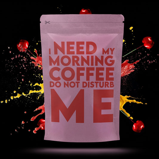 Roze koffiezak met grote rode tekst: "I need my morning coffee. Do not disturb me", geplaatst tegen een achtergrond met spattend rood en geel sap en vliegende kersen.